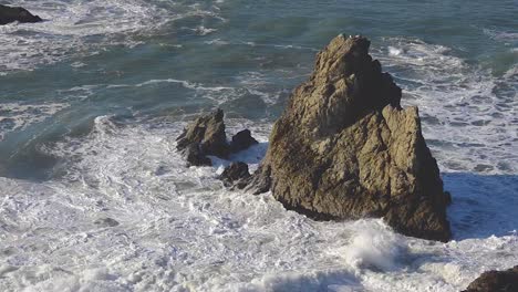 powerful pacific ocean waves crashing against a large ocean sea stack rock formation with birds sitting on top of it - san francisco, california