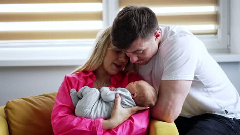 blonde woman in pink shirt holding tiny newborn. father is sitting beside soothing his little child.