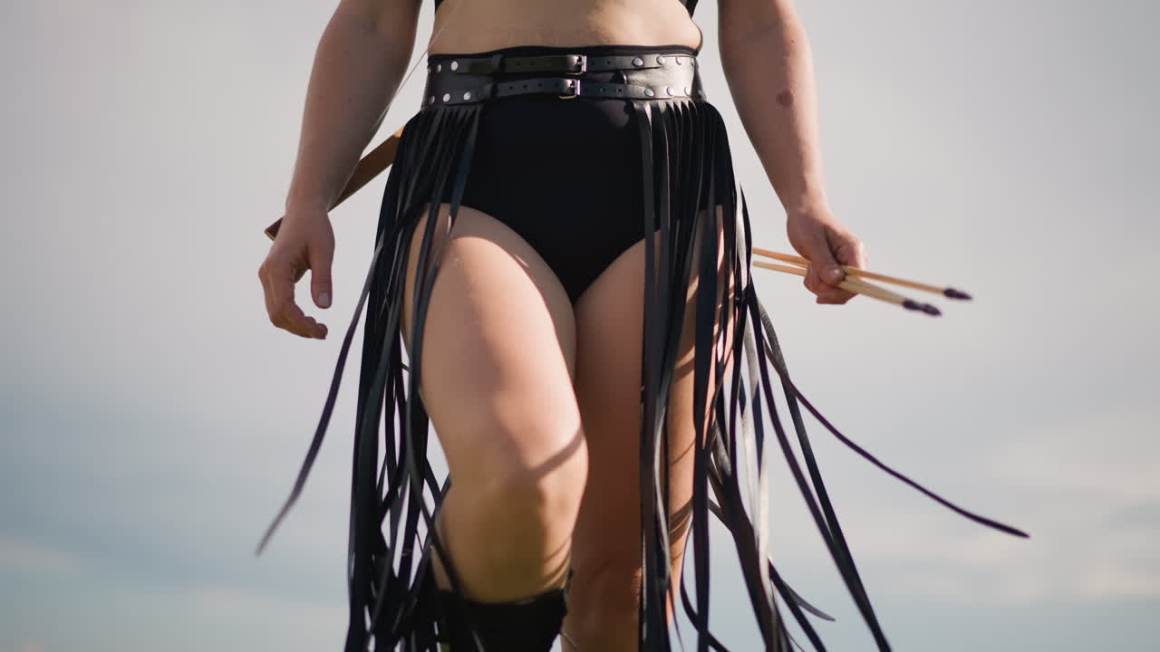 Inquadratura frontale di un batterista con gonna a frange e cintura, passo sicuro con le bacchette, aria da leader di una parata e artista di strada, orizzonte illuminato dal sole e sfondo di spiaggia estiva, movimento ritmico e festival