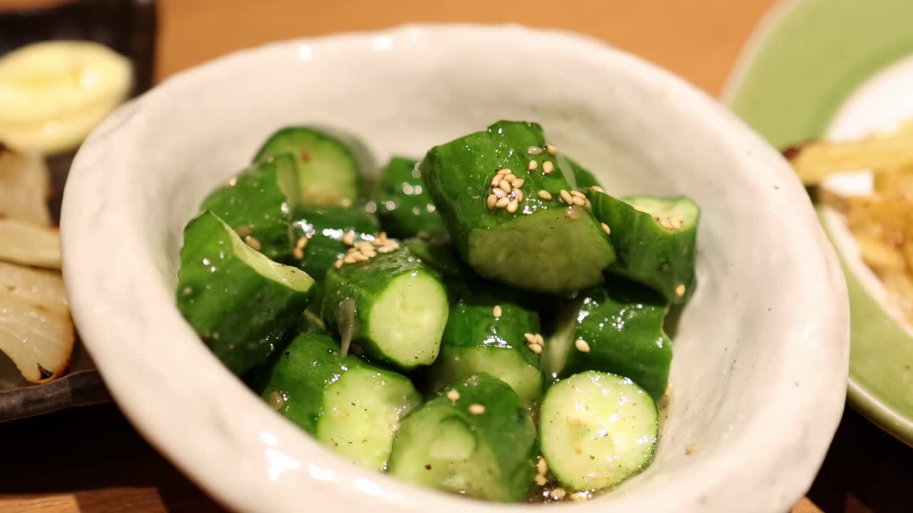 Close-up of chopsticks stirring sliced cucumbers with sesame seeds in a white bowl on a dining table.
