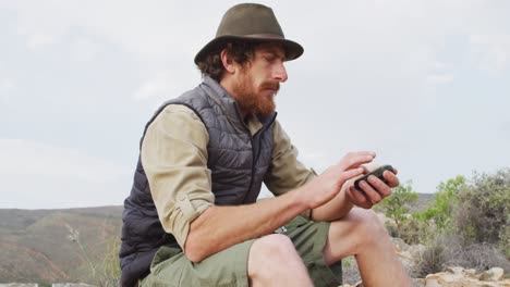 Bearded-caucasian-male-survivalist-sitting-on-mountain,-holding-compass-and-looking-around
