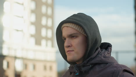 atleta enfocado en sudadera y gorra estirando el brazo contra un fondo urbano brillante, mostrando determinación y preparación para la actividad física, edificios modernos iluminados por el sol