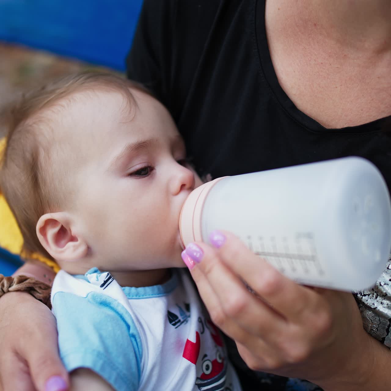 Baby nutrition during the walk outdoors. Sweet boy suckling milk from bottle and rubbing his foot. Close up. Blurred backdrop