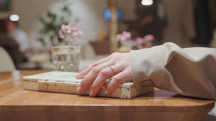 Nahaufnahme einer Frauenhand, die bei sanfter Innenbeleuchtung nach einer alten Farbpalette auf einem Holztisch greift und sie aufhebt. Die Szene umfasst verschwommene Hintergrundelemente wie Menschen, Pflanzen und Blumen.
