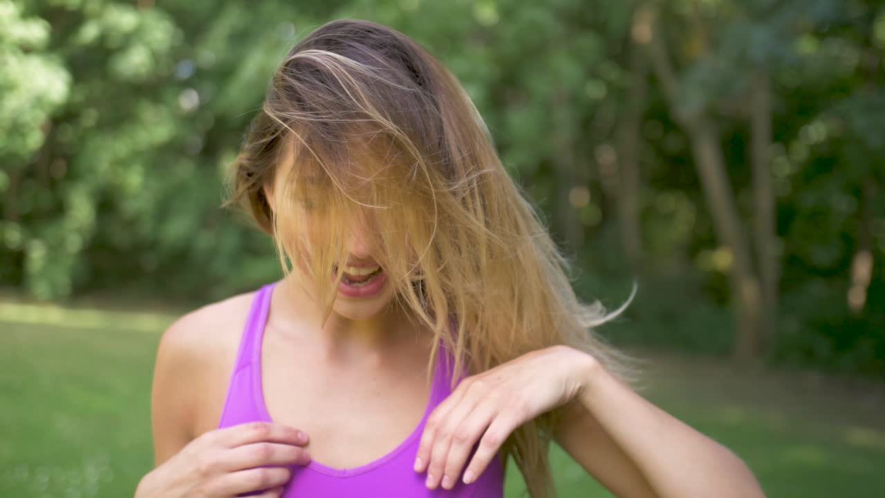 una mujer con un top deportivo morado sonríe, se sacude el pelo y gira la cabeza posando para la cámara.
