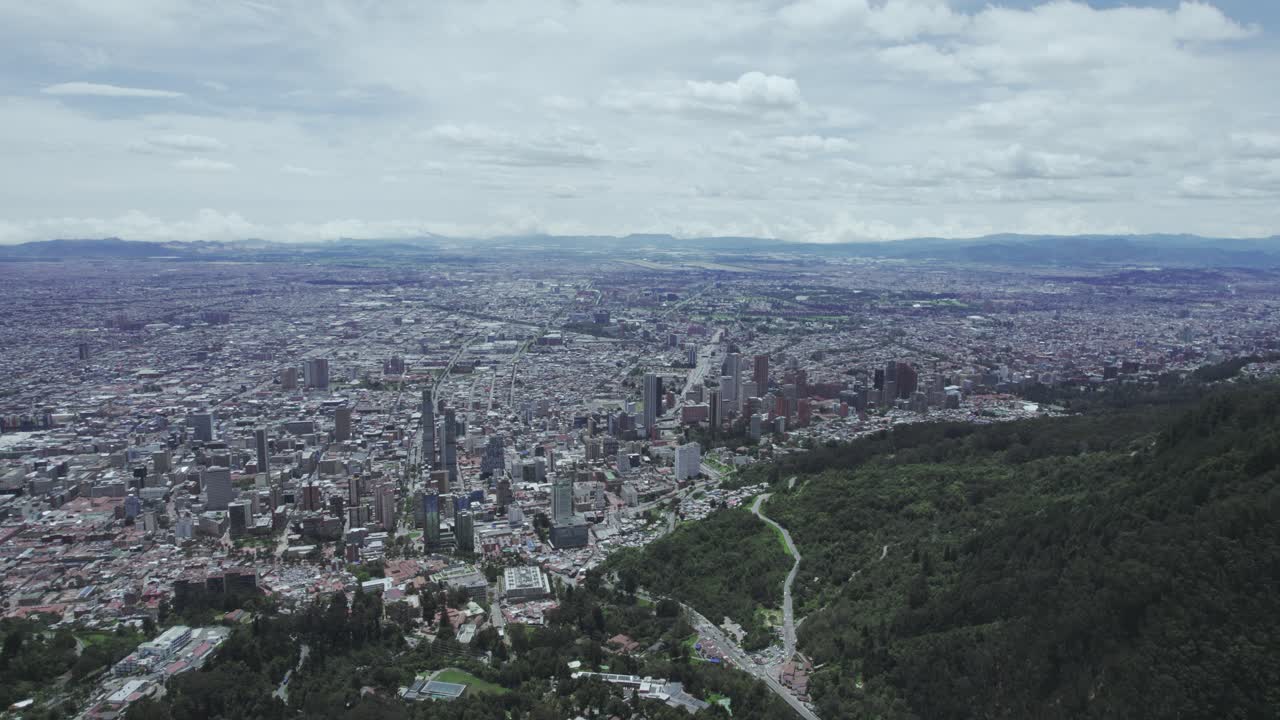 Aerial view from the lush green mountain top to the vast city of Bogotá, with a close-up in direction to the clouds filling the sky.