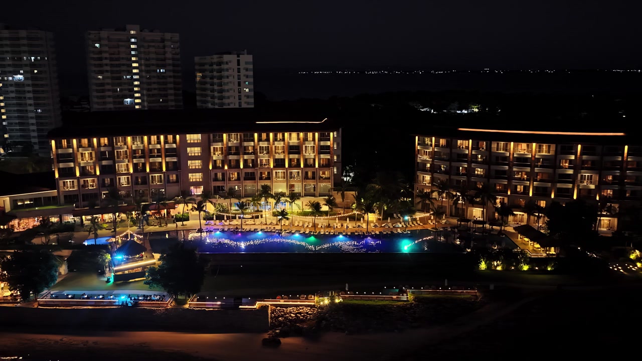 Uno scatto aereo notturno del resort Dusit Thani Mactan Cebu nelle Filippine, che evidenzia l'area della piscina illuminata e gli edifici circostanti che si riflettono sull'acqua.