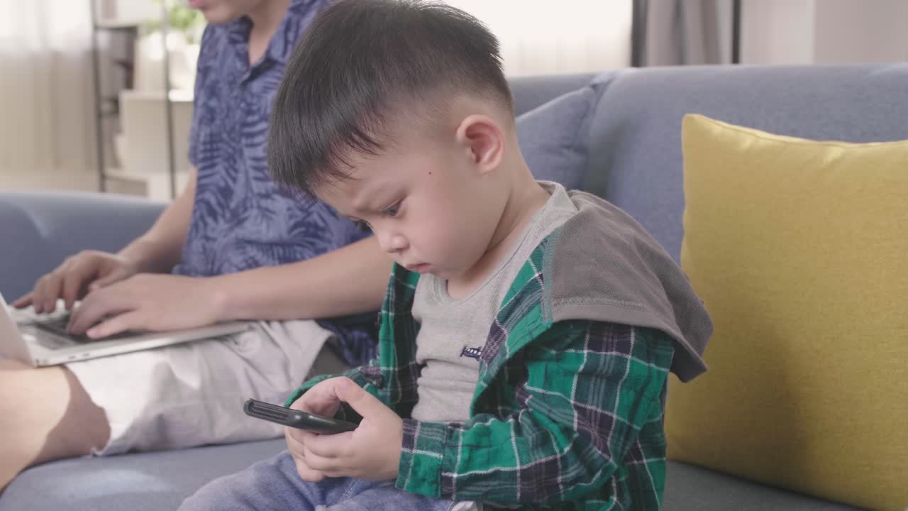 Asian Boy Using Smartphone While Father Working On Laptop