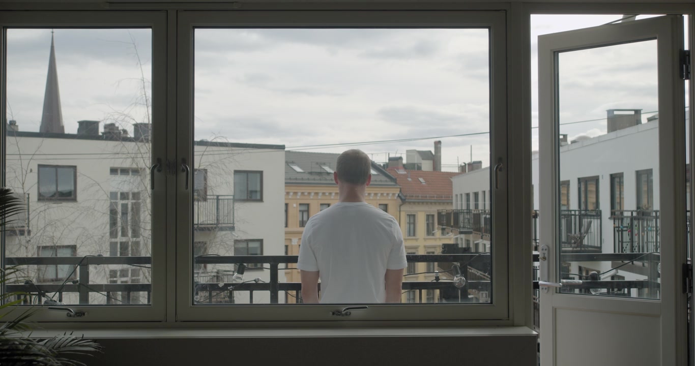 Toma de cámara de cine estática trasera y amplia de un hombre noruego aislado y solo en un apartamento oscuro en cuarentena por Corona, de pie en el balcón, con el fondo de la ciudad, en la parte central de Oslo, Noruega