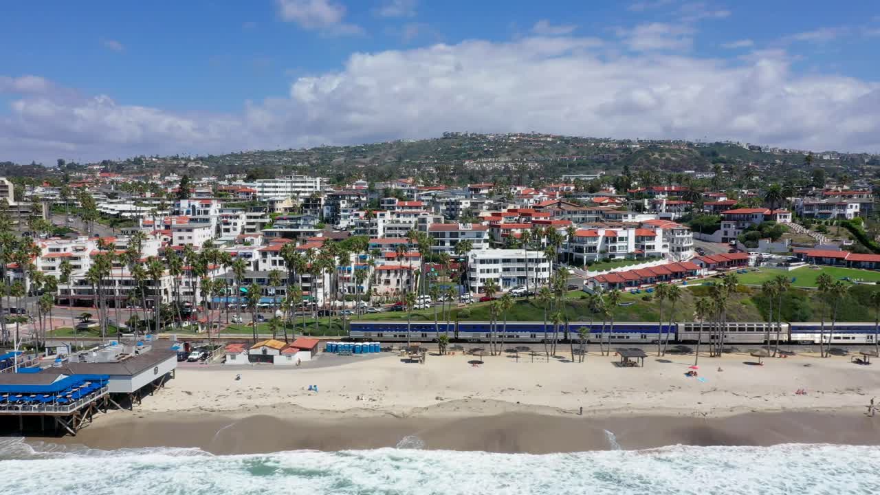 4k Aerial Video, left-to-right showing train passing San Clemente Pier and beach and shops in Orange County, towards San Diego, California,