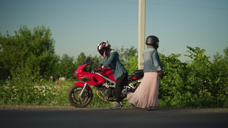 two women wearing helmets, one seated on a red sports bike while the other drops off the bike, they are on a roadside, with cars passing by in the background