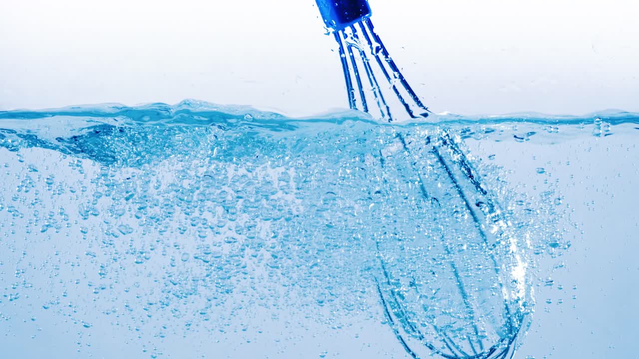 Batidor de metal agitando agua y creando salpicaduras dinámicas con burbujas de aire en un líquido azul claro sobre un fondo claro.