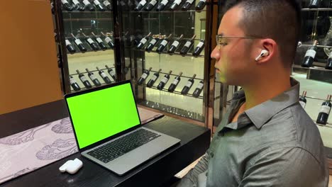 Wealthy-Asian-Male-Wearing-Earbuds-and-Enjoying-Music-In-Front-of-Laptop-with-Green-Screen,-Sitting-Inside-Luxurious-Home