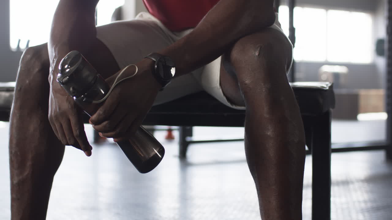 Resting on bench, man holding water bottle and wearing fitness tracker in gym