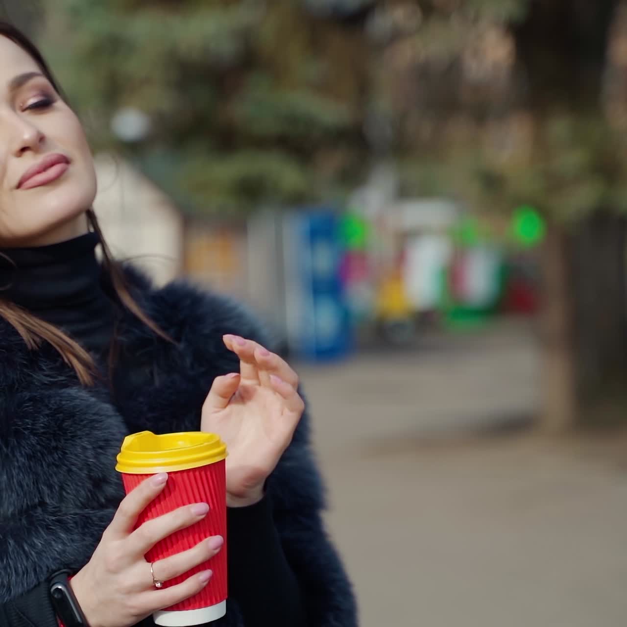 Lovely girl in winter coat. Beautiful young woman holds a plastic cup of coffee and smiles on camera in the park. Slow motion.