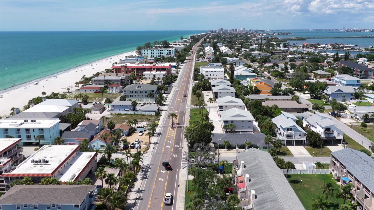 Kustvägen löper mellan strandnära lägenheter och turkosa golfvatten i Indian Rocks Beach, Florida. Bilar passerar den vita sandstranden nära Clearwaters avlägsna horisont. Flygfoto med drönare, sidled vidvinkel.