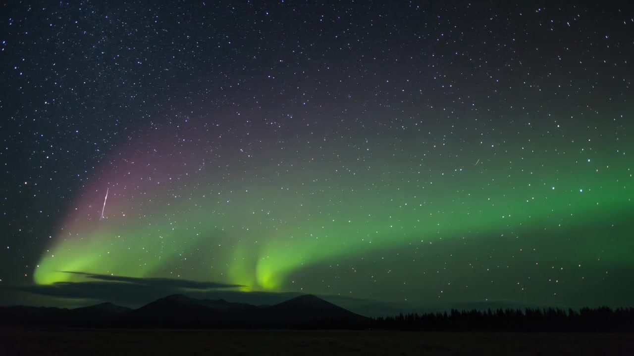 Vento solare che alimenta l'aurora da una fascia verde a due picchi sopra il bordo polare, con stelle e una meteora.