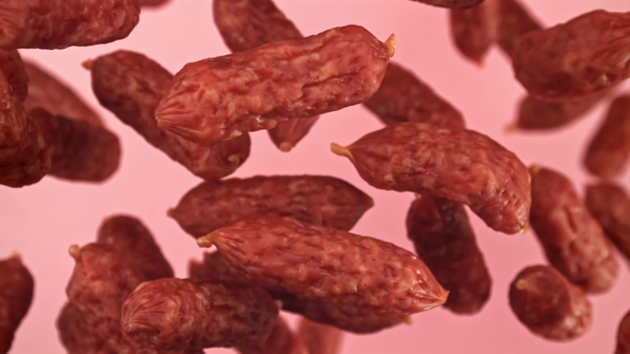 Mini kiełbaski salami spadające w zwolnionym tempie na różowym tle.