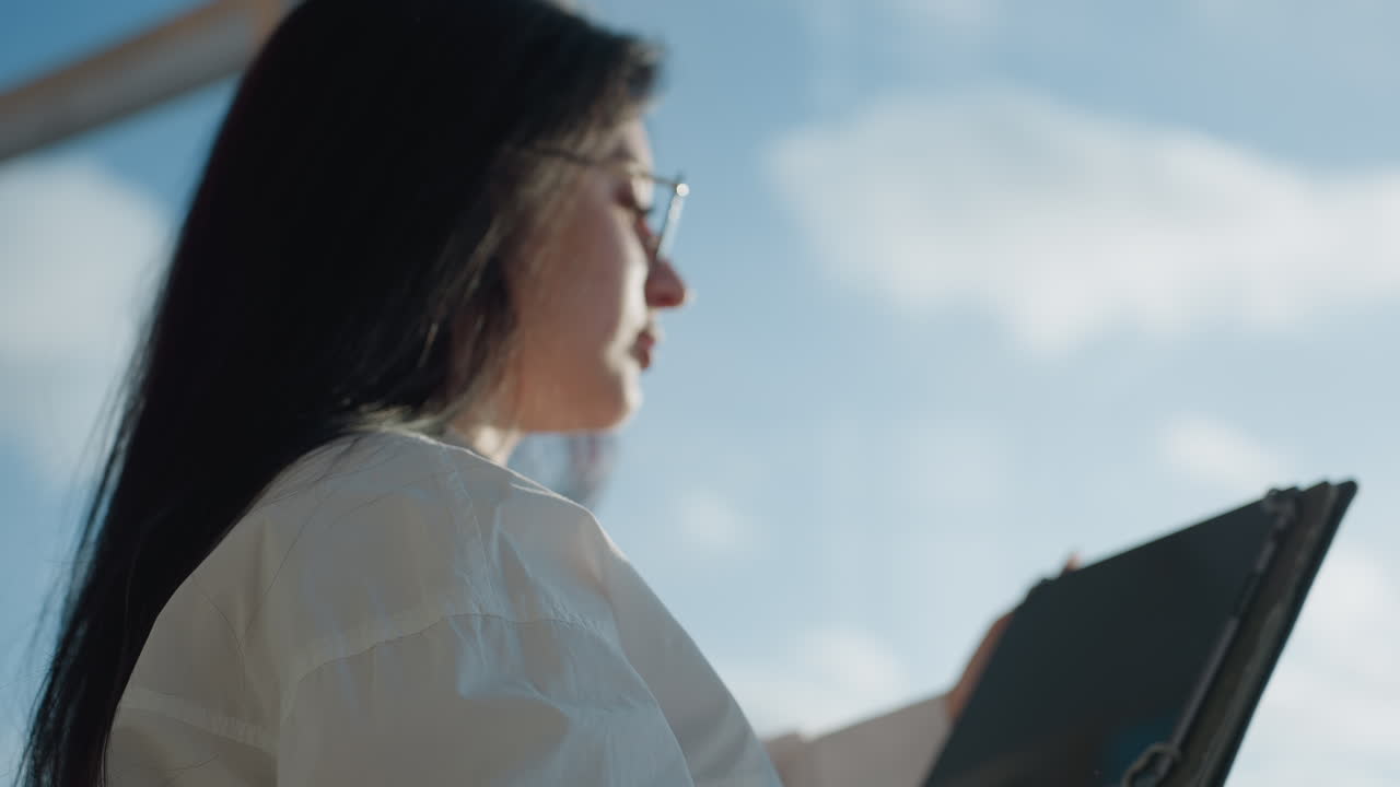 Rear view of woman in white shirt adjusting glasses while using tablet against soft blurred background of clouds and sky, bathed in natural sunlight and framed with a relaxed tech focused vibe