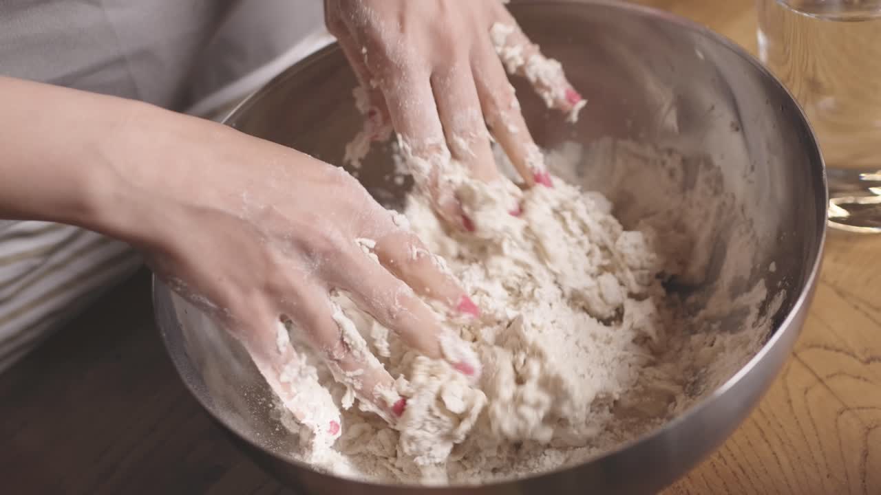 Close up of a baker's hands pouring water into a stainless steel bowl to mix organic flour into dough on a rustic wooden table