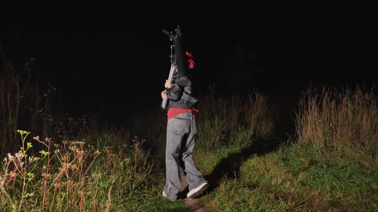 une personne installant un télescope de nuit, un sweat à capuche rouge ajustant le trépied et le télescope sur un chemin herbeux, se préparant à l’observation des étoiles et à l’astrophotographie, atmosphère rurale paisible, ciel sombre en arrière-plan, concentré