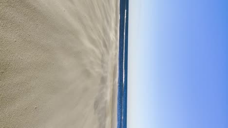 La-Arena-De-La-Playa-Es-Volada-Por-El-Viento,-La-Brisa-De-Verano,-El-Video-Vertical-De-Un-Cielo-Azul-Brillante-De-Ensueño,-Una-Tormenta-De-Arena-Amistosa-Con-La-Playa