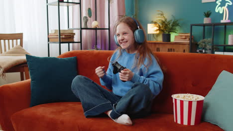 girl child using joystick controller playing video console television game sits on sofa in room