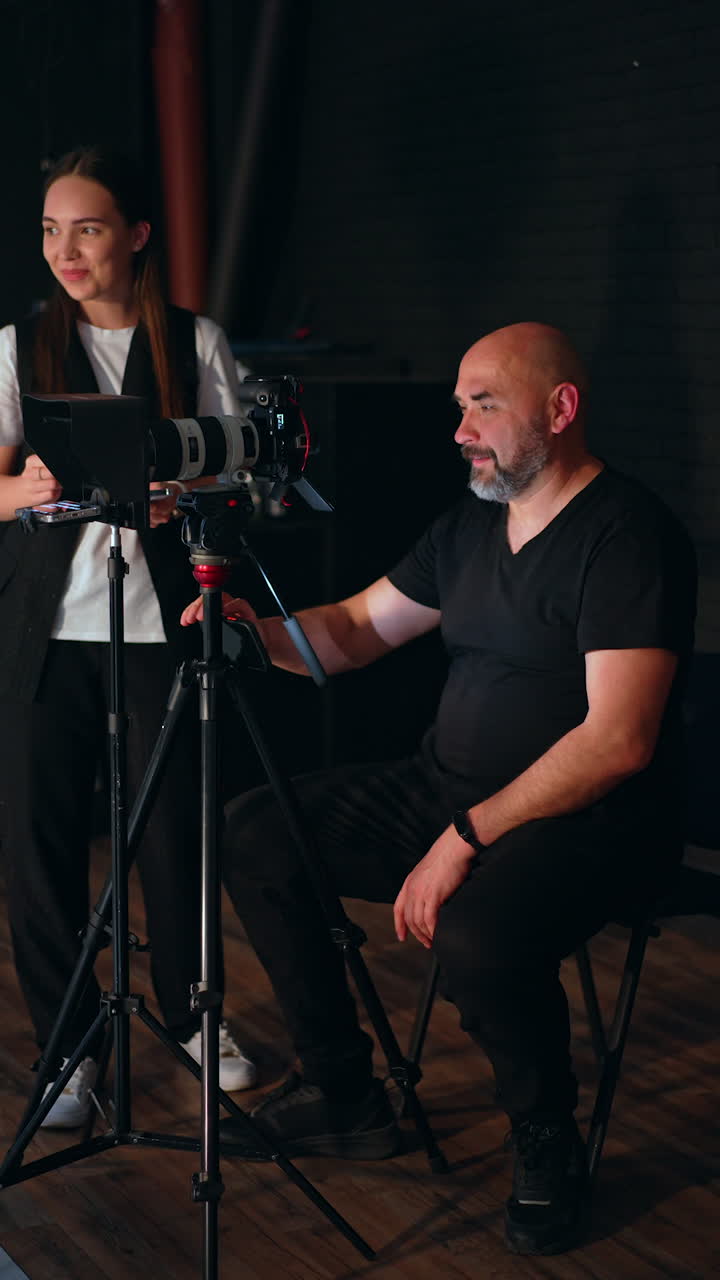 Professional photographer sits behind his camera on tripod. Positive smiling brunette stands behind. Studio footage. Vertical video.