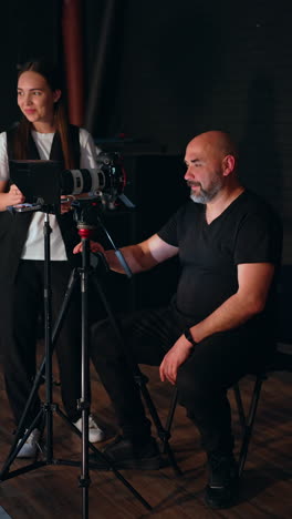 professional photographer sits behind his camera on tripod. positive smiling brunette stands behind. studio footage. vertical video.