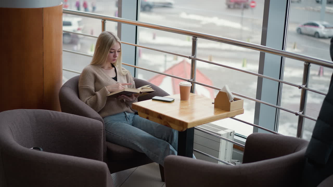 joven sentada cómodamente en un acogedor café, absorta en la lectura de un libro con una taza de café en el techo de la mesa, mirando desde una gran ventana, con una vista urbana al aire libre nevada a través de un panel de vidrio