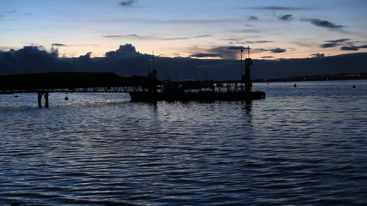 Black pier in a blue water marine bay; water looks cool and fresh, small waves reflect gray autumn light. No one is around. Sun has set. Footage in HD