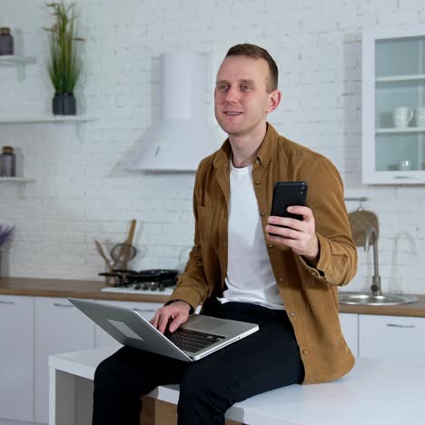 portrait of a student with a laptop and a phone. smiling young man is sitting on a kitchen table and using a laptop and mobile phone at home. quarantine concept