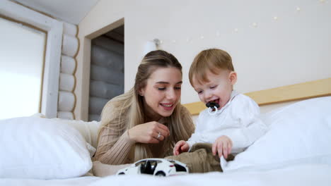 mother and little toddler baby sit on the bed. kid with pacifier plays with a white car. low angle view.