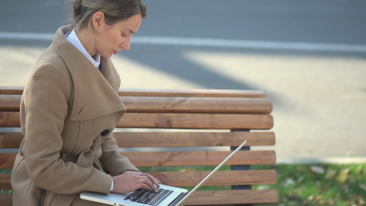 Woman in a brown coat working on her laptop on a bench in the park