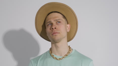 joven modelo posando con un gran sombrero marrón, collar dorado y camiseta verde y tocándose la barbilla