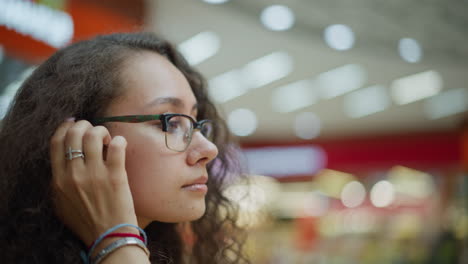 young woman with curly hair standing thoughtfully in well-lit mall, adjusting her hair with a gentle expression, the background features soft bokeh lighting