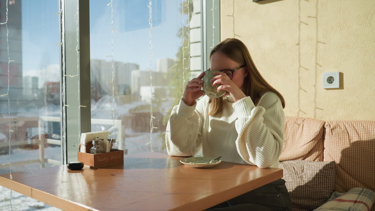 Side view of woman in white sweater holding green tea cup near window in cozy cafe, sunlight highlighting her face, snow covered street and tall buildings visible through decorative glass panel
