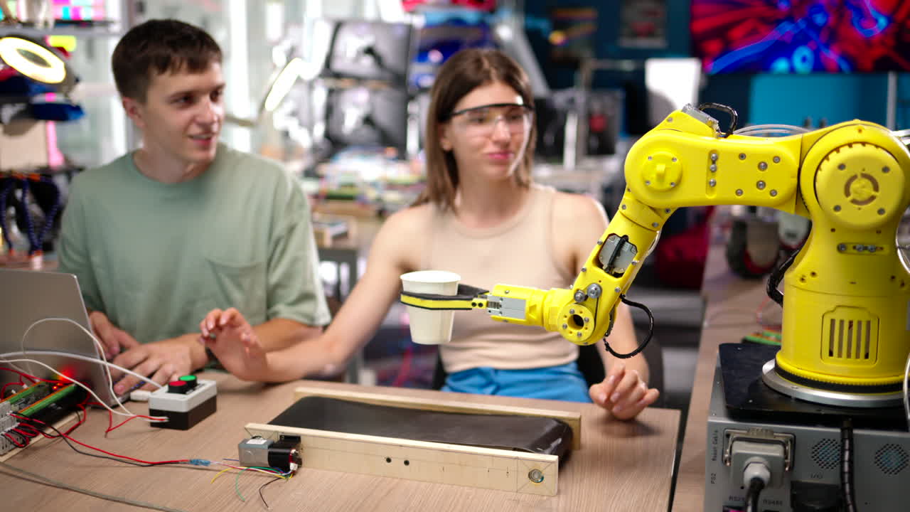 Young happy engineers programming an yellow robotic arm in the workshop to grab cardboard water glass, computer programming training for coffee preparation, celebrating success