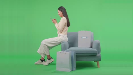 vista lateral de una mujer asiática de compras con bolsas de compras usando teléfonos móviles compras en línea girarse para sonreír mientras está sentada en el sofá frente a la pantalla verde