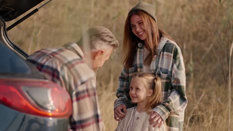 Close-up-shot-of-a-happy-brunette-girl-in-a-green-plaid-shirt-hugging-her-little-daughter-and-communicating-with-her-husband,-a-middle-aged-man-who-is-sitting-in-the-open-trunk-of-a-black-car-during-their-picnic-outside-the-city-in-the-summer