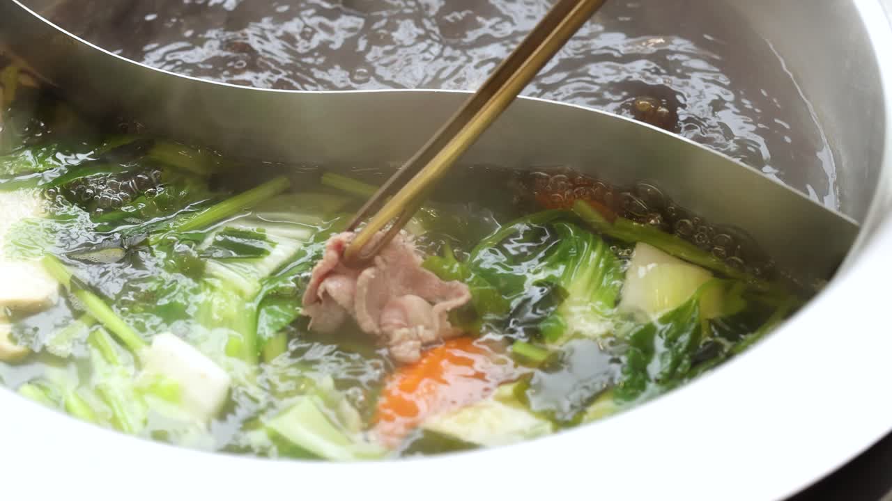 Chopsticks cook beef slice in steaming dual broth hotpot with assorted vegetables, overhead view