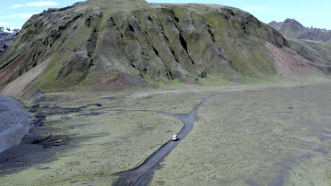 Aerial-of-4x4-car-driving-on-a-black-sandy-mountain-road-in-front-of-rocky-mountains-covered-by-a-green-moss