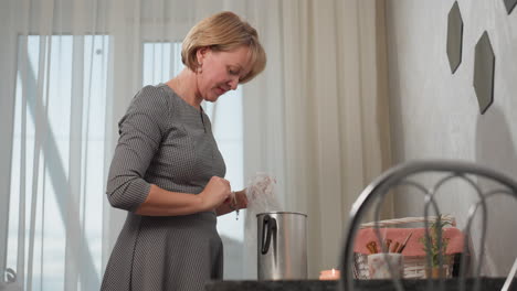 young woman in patterned dress thoughtfully drops translucent leather-like material into metal jug while standing at table with creative supplies, candle, and basket near bright window