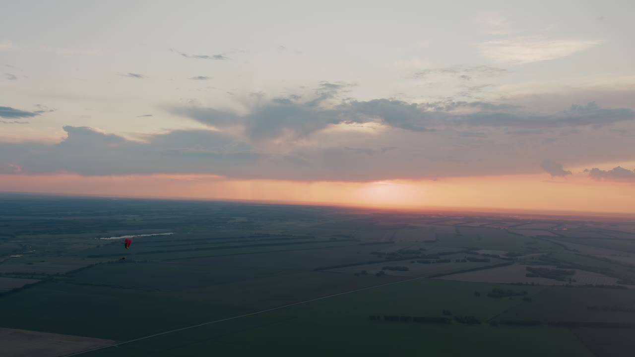 Distant silhouette of pilot gliding beneath red canopy across pastel sunset sky, clouds drifting above sprawling patchwork fields and tree lines