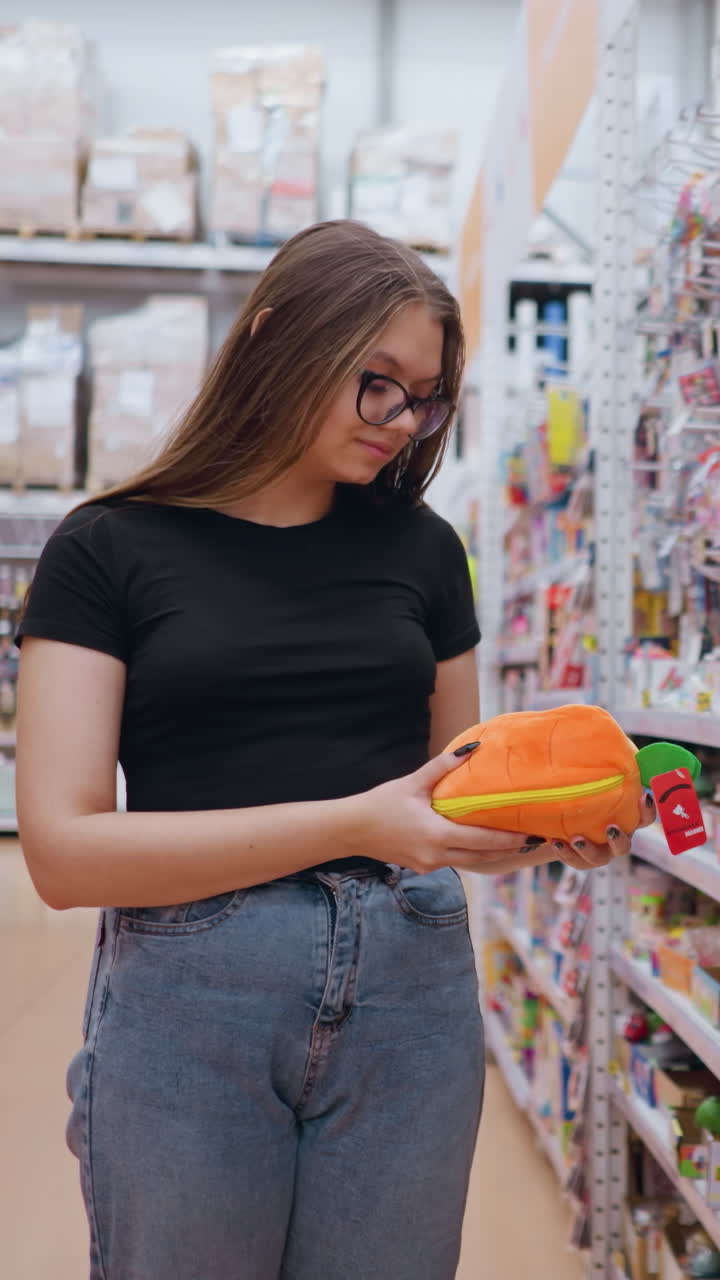 mujer joven en camiseta negra y vaqueros recogiendo una bolsa de peluche naranja brillante de un estante en una tienda de juguetes bien iluminada, juguetes coloridos y accesorios llenan los estantes mientras navega por las opciones
