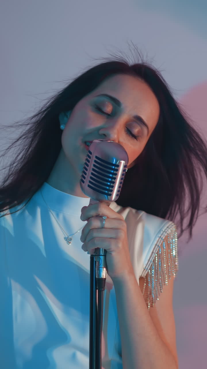 una cantante con un vestido blanco canta apasionadamente, sosteniendo un micrófono vintage con una mano contra un fondo azul. su cabello fluye hacia atrás