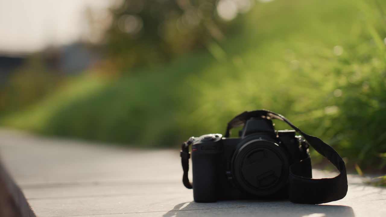 กล้องวางอยู่บนขอบถนนใกล้หญ้า กล้อง DSLR สีดำวางอยู่ข้างเป้สะพายหลังบนทางเท้าที่แสงแดดส่องถึง ระยะชัดตื้นและโบเก้ที่นุ่มนวล มือเอื้อมไปหยิบกล้อง ช่างภาพสมัครเล่นกำลังหยุดพักในช่วงแสงสีทอง