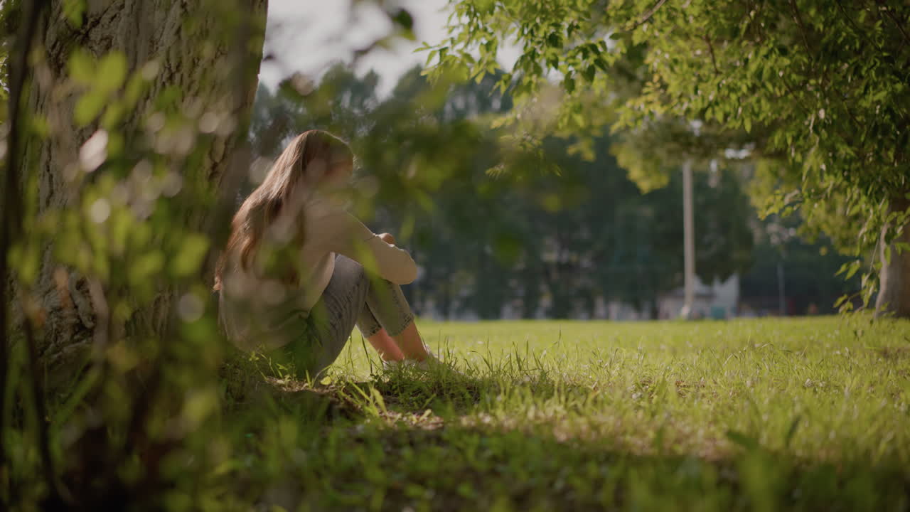 잔디에 앉아있는 여자의 뒤쪽 풍경, 그녀의 무 위에 팔을 접고 생각에 빠져, 빛이 부드럽게 빛나고, 조용한 설정에는 흐릿한 나무, 경기장 기둥이 포함됩니다.