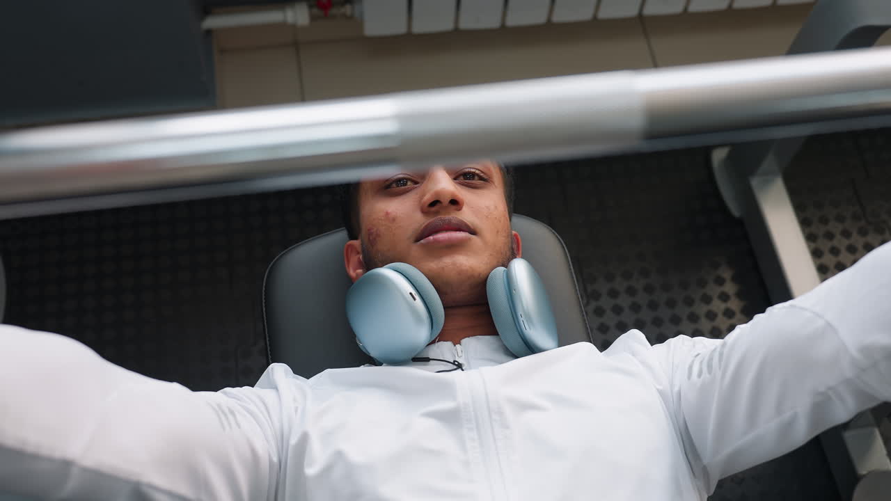 top down view of man in white jacket with wireless headphones around neck lying on bench press station pressing barbell with controlled form muscle tension visible gym interior background