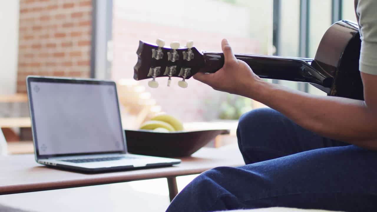 African american man plays guitar and singing, using laptop at home